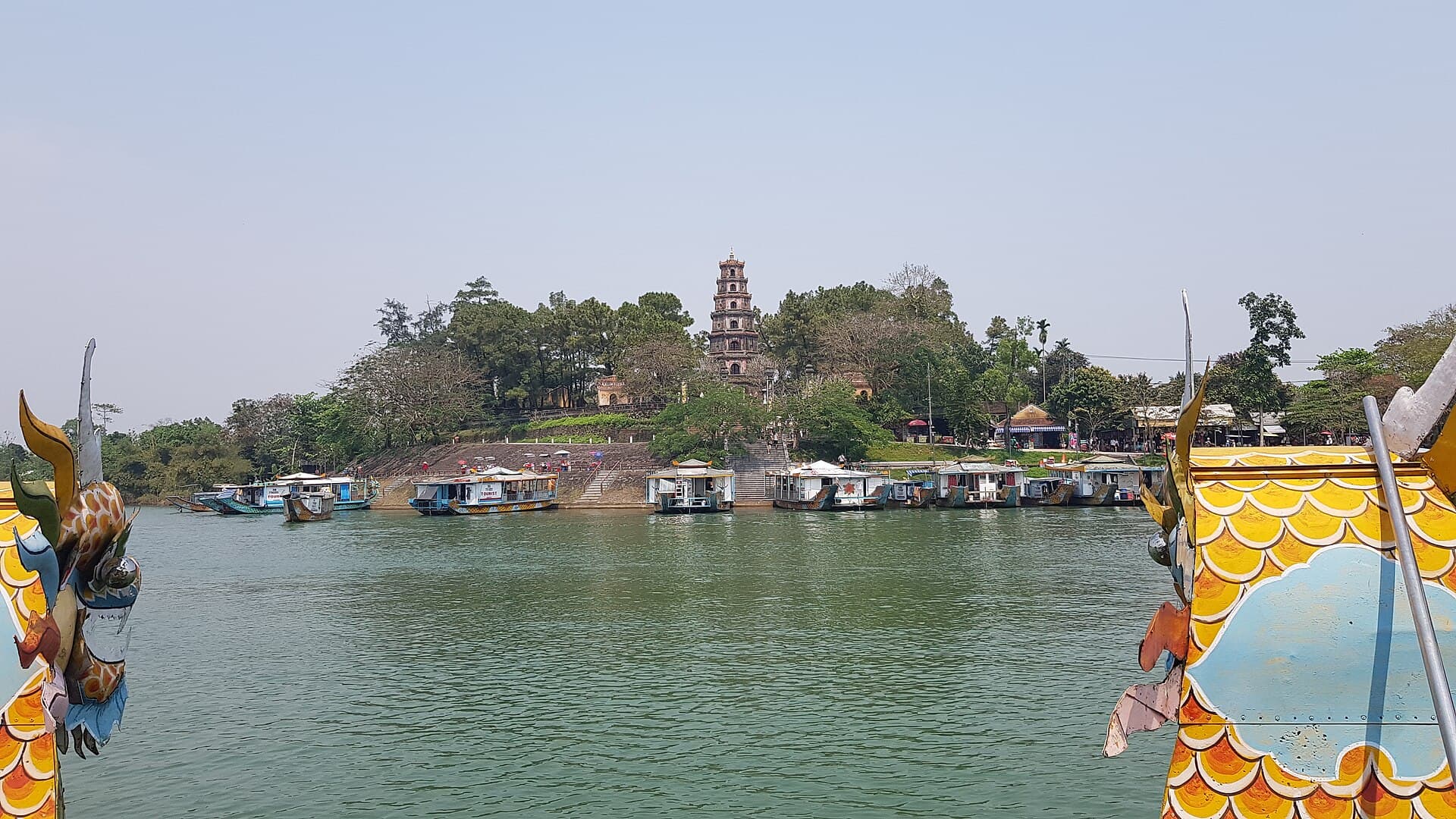 Thien Mu Pagoda's seven-tier Phuoc Duyen tower rising above the trees on the north bank of the Perfume River in Hue, a row of dragon boats moored at the steps below and the painted dragon-head prows of another boat framing the view