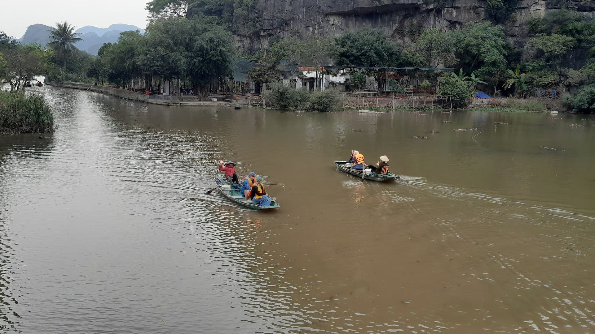 Two small boats crossing the still water of the Van Long nature reserve in Ninh Binh, with a village and limestone cliffs behind