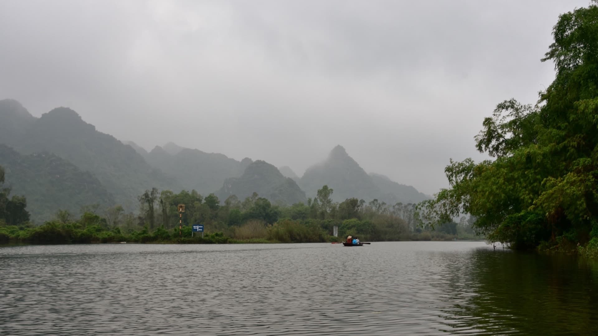 A single hand-rowed boat with a conical-hatted rower crossing the misty Yen stream toward the Perfume Pagoda, with limestone hills fading into low cloud behind